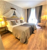 Ett hotellrum med två sängar, en himmelssäng, en sittgrupp med en fåtölj och ett stort fönster med gardiner.