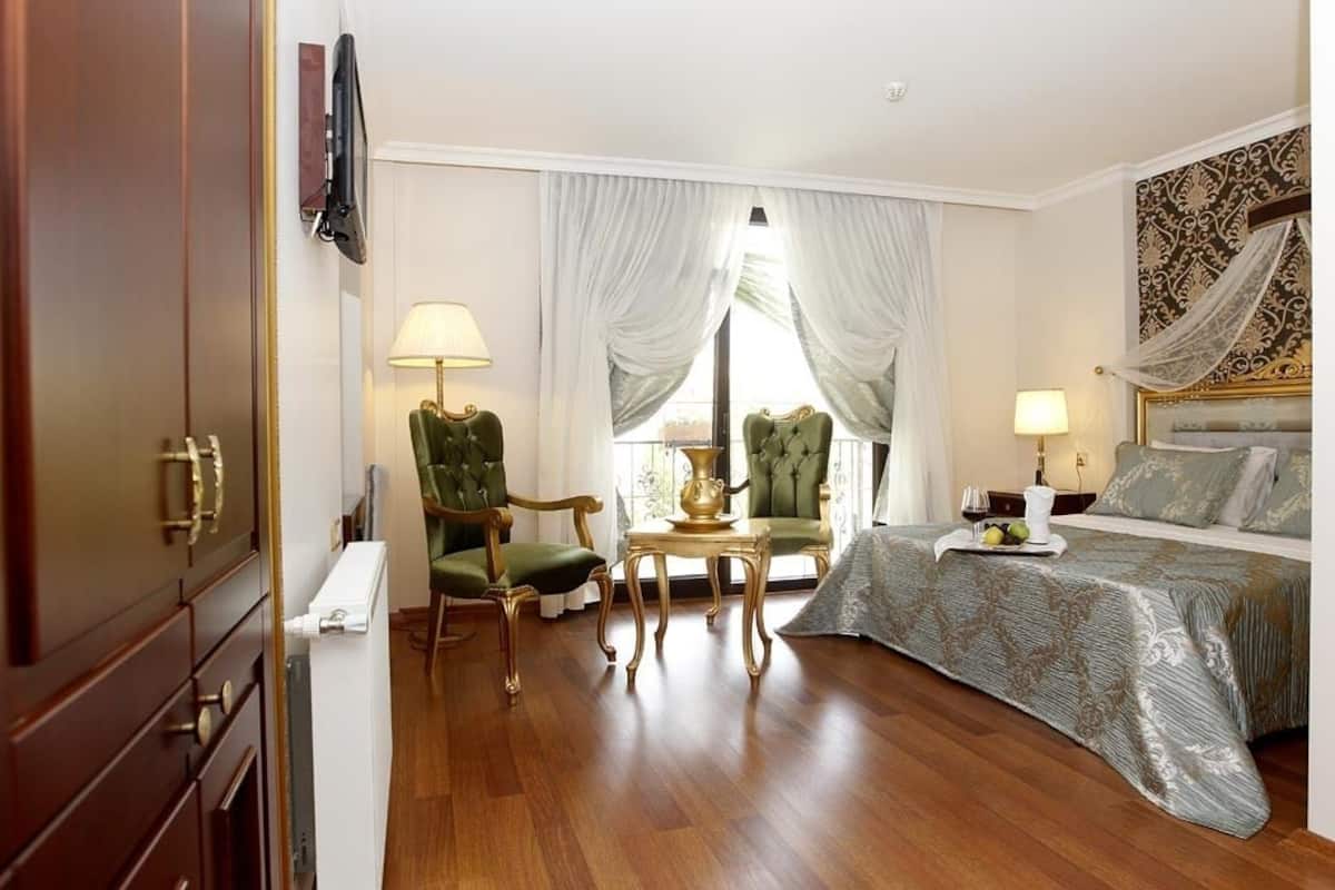 A hotel room with a bed, two green armchairs, a small table, a lamp, and a television mounted on the wall.