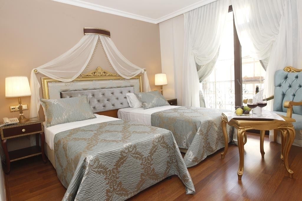 Quarto de hotel com duas camas, incluindo uma cama de dossel, um espaço de estar com uma poltrona e uma mesa com um vaso e frutas.