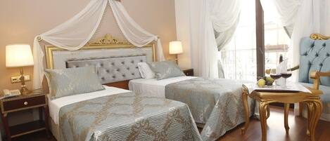 Quarto de hotel com duas camas, incluindo uma cama de dossel, um espaço de estar com uma poltrona e uma mesa com um vaso e frutas.