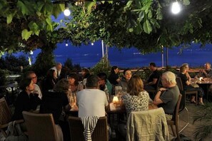 Un espace de restauration en plein air, avec des tables et des chaises disposées sous une canopée d’arbres, offrant une vue sur une étendue d’eau.