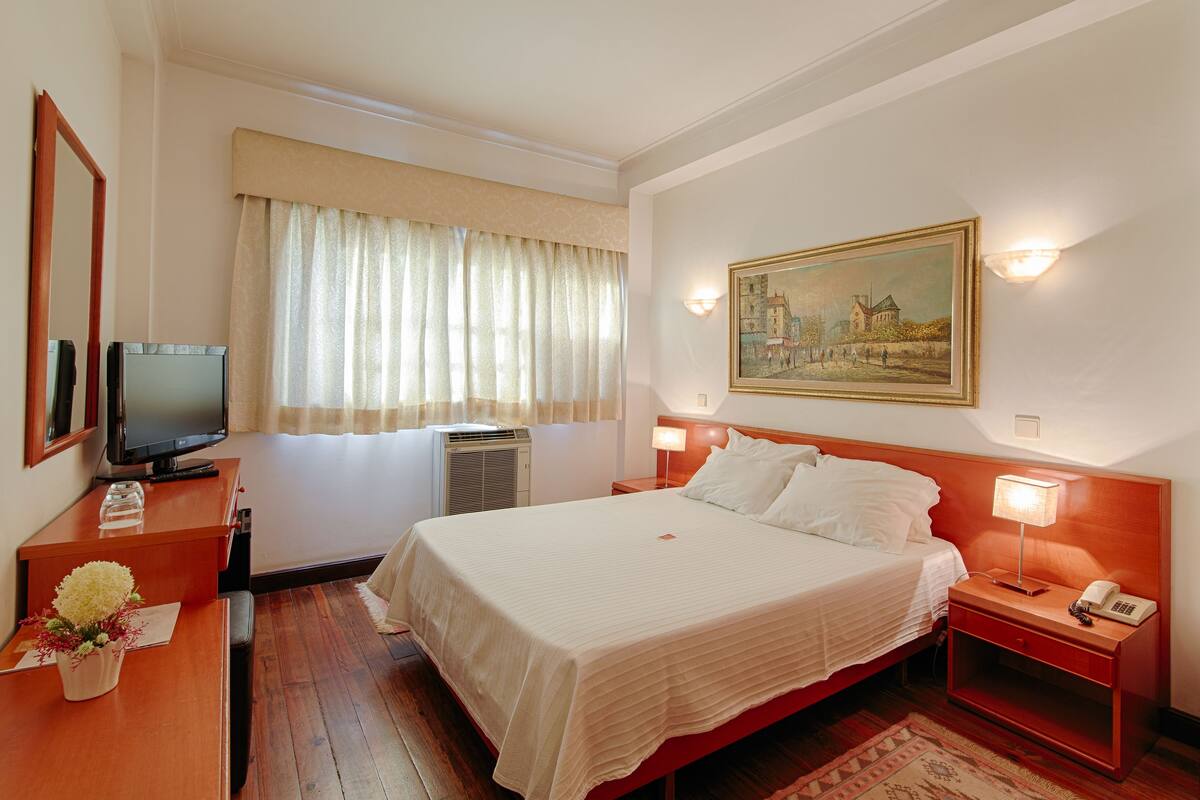 A hotel room with a large bed, a television, a desk with a vase of flowers, and a painting on the wall.