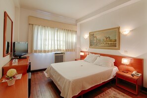 Una camera d'albergo con un letto grande, una televisione, una scrivania con un vaso di fiori e un quadro appeso al muro.