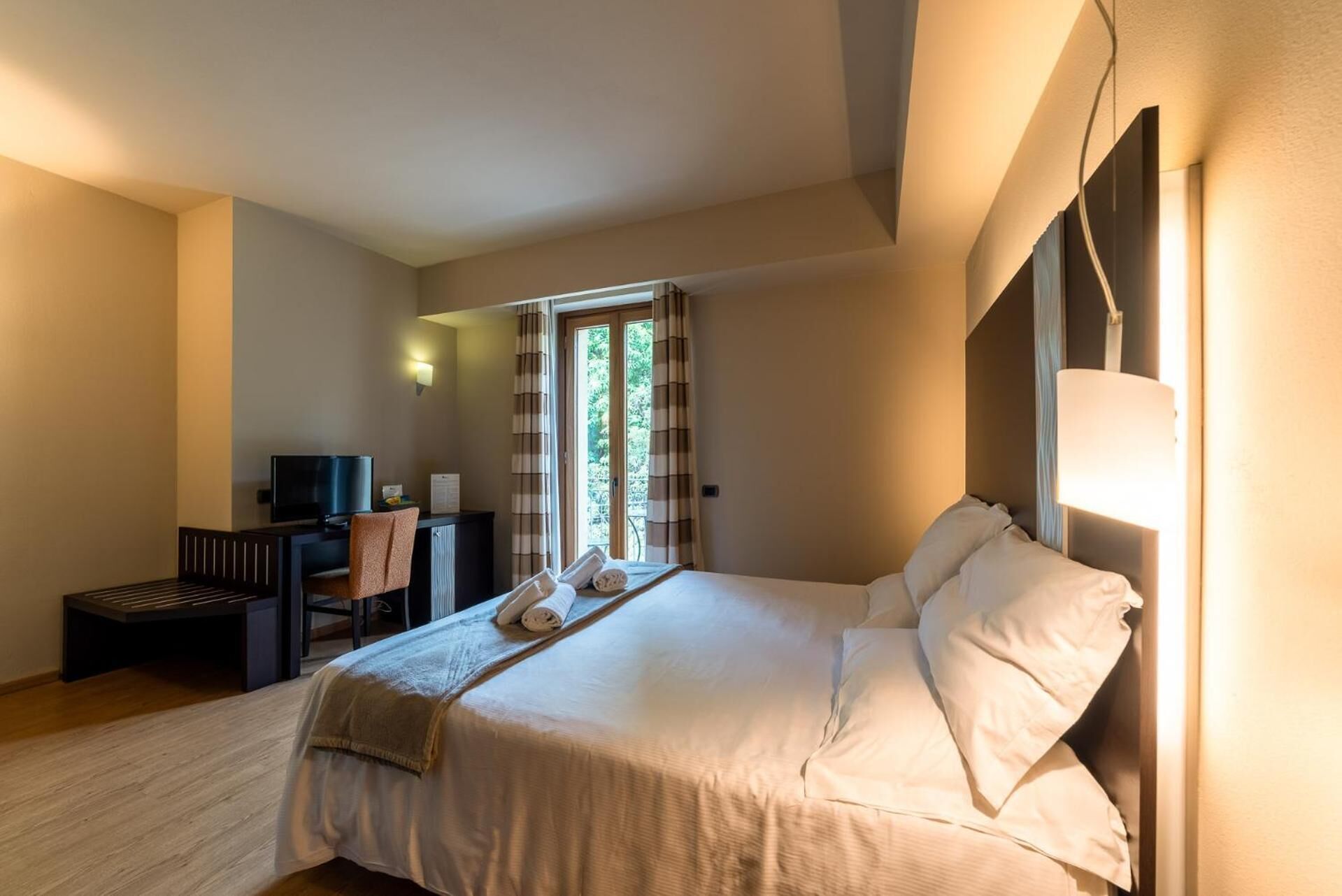 A hotel room with a bed, a desk with a chair, a television, and a window with curtains.