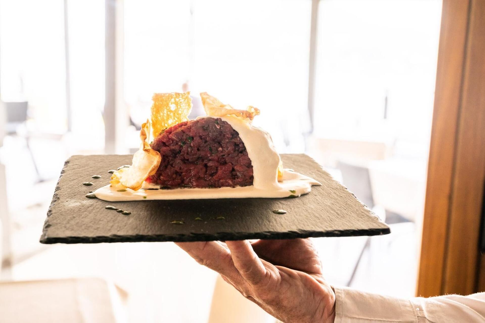 A person holding a black slate serving board with a dish of meat and sauce, and a piece of bread.