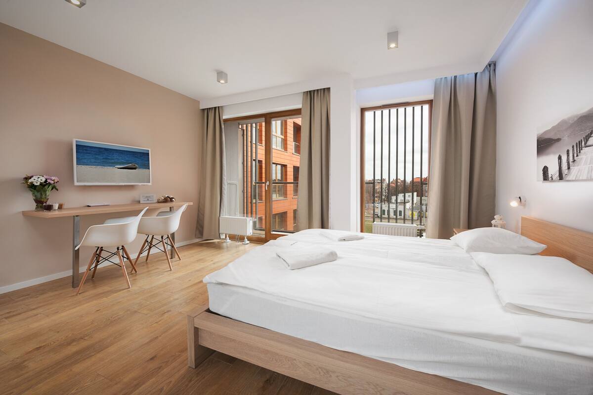 A modern hotel room with a large bed, a desk with chairs, a television, and a view of the city through large windows.