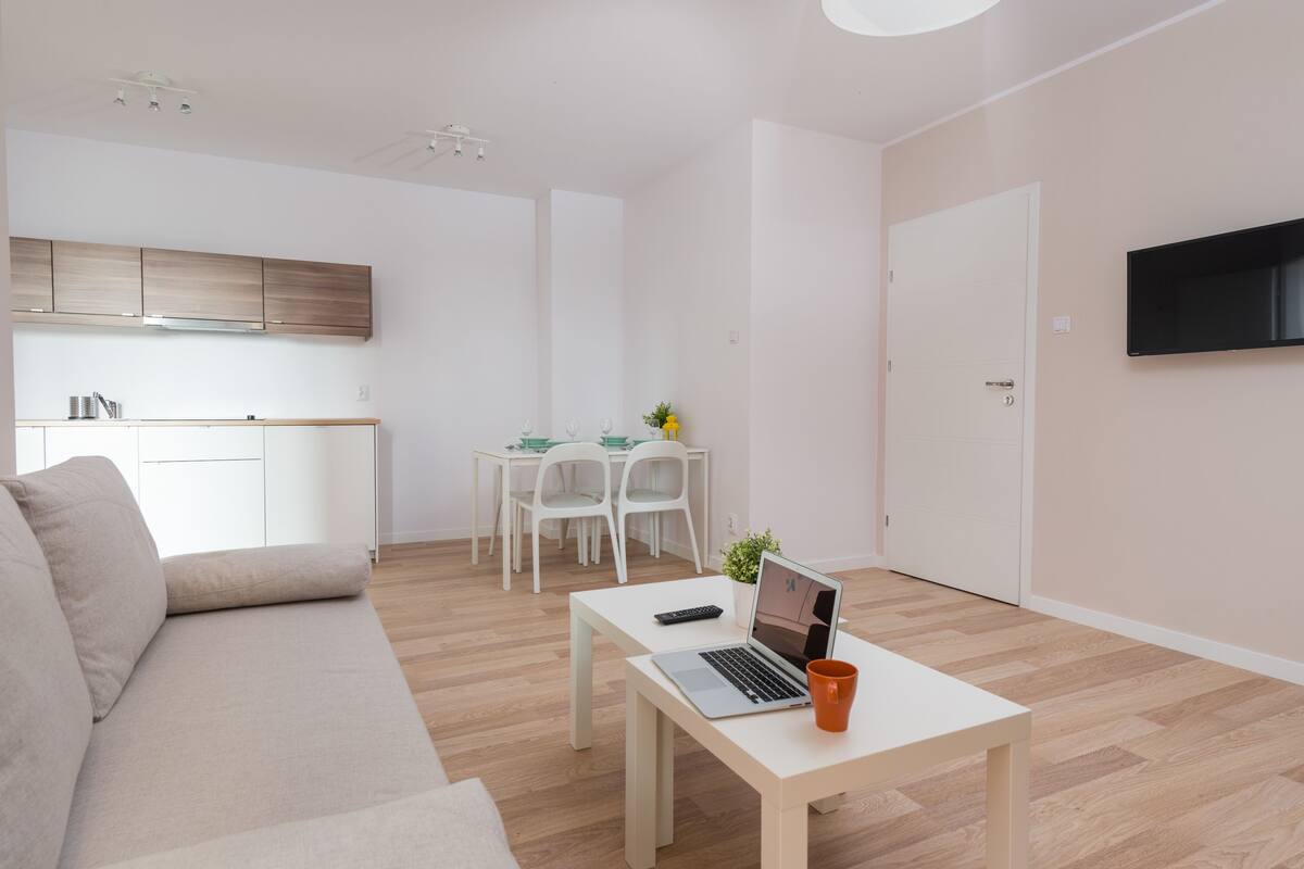A modern, minimalist living room with a sofa, a coffee table with a laptop and mug, a TV mounted on the wall, and a kitchen area in the background.