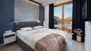 A modern bedroom with a large bed, a wall-mounted light, a window with curtains, and a view of the outside. - Apartinfo Aura Apartments (Gdansk)