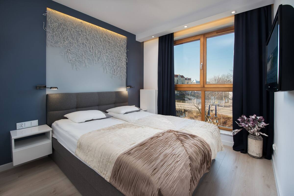 A modern bedroom with a large bed, a wall-mounted light, a window with curtains, and a view of the outside.