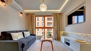 A modern living room with a sofa, a glass-top coffee table, a TV mounted on the wall, and a large window with curtains. - Apartinfo Aura Apartments (Gdansk)