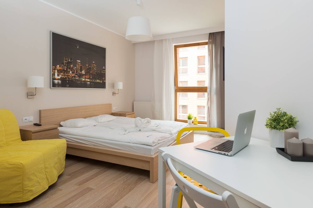 A modern hotel room with a bed, a desk with a laptop, a yellow chair, a cityscape painting, and a window with curtains.