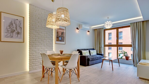 A modern living room with a dining area, featuring a brick wall, white furniture, and large windows. - Apartinfo Aura Apartments (Gdansk)