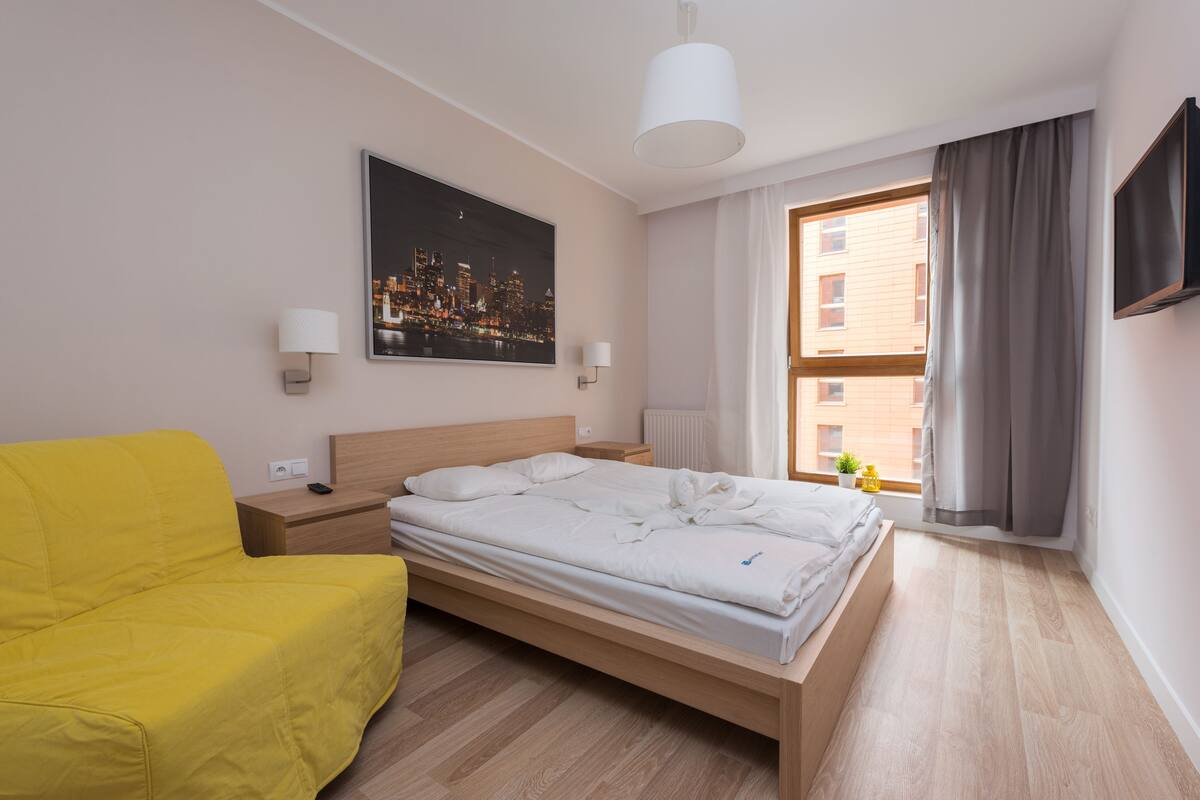 A hotel room with a bed, a yellow armchair, a cityscape painting, a window with curtains, and a wall-mounted TV.