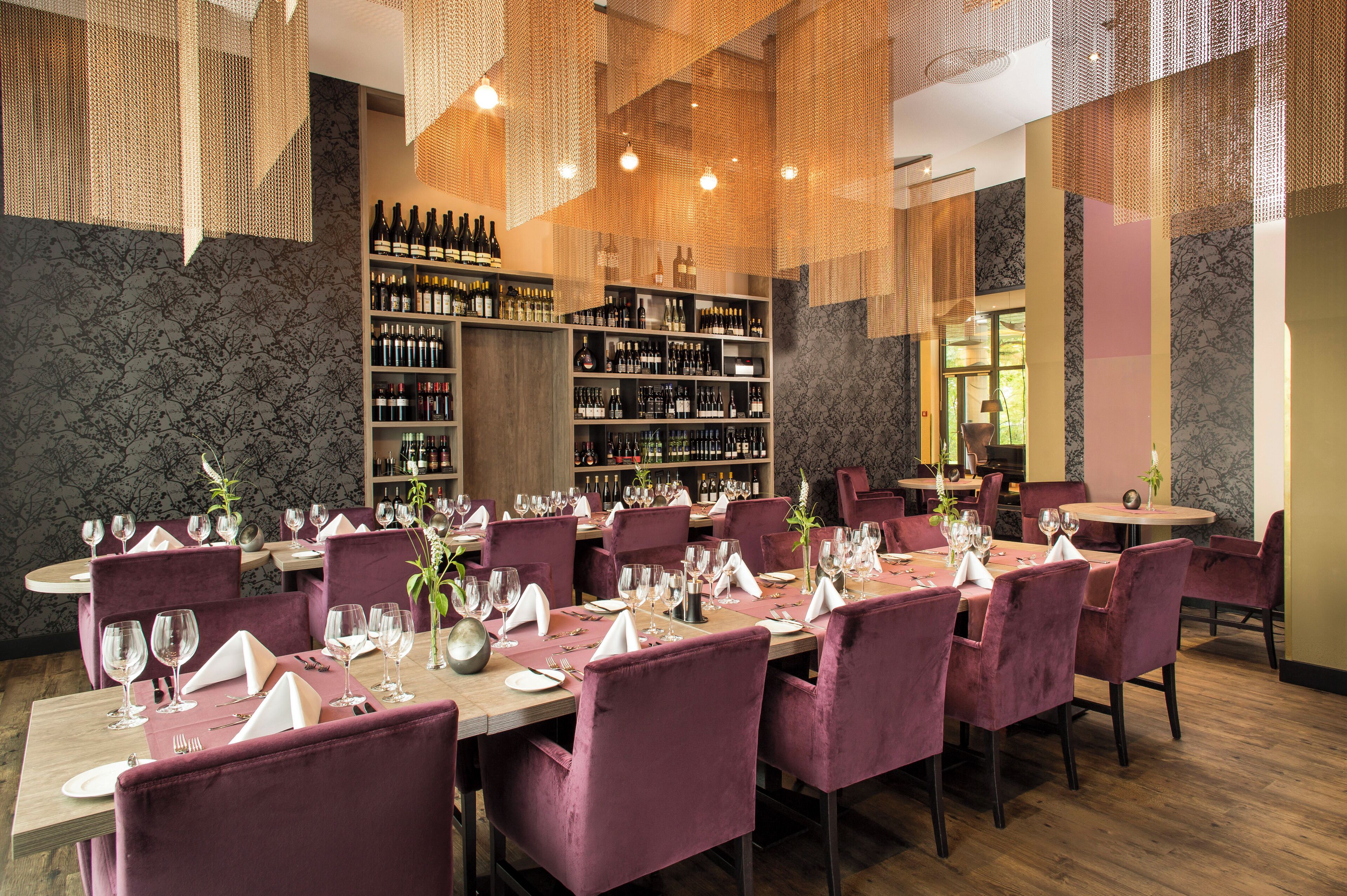 Ein Restaurant mit mehreren Esstischen, gedeckt mit Weingläsern, Tellern und Besteck. Regale mit Weinflaschen und ein großes Fenster, das für natürliches Licht sorgt.