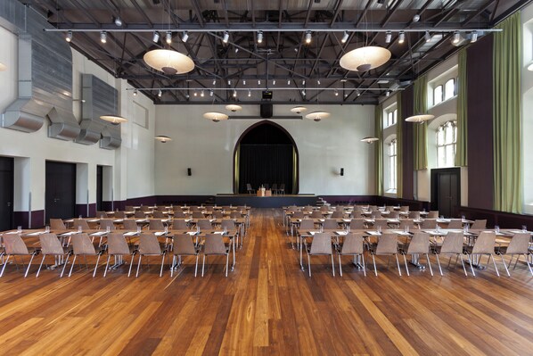 A large hall with rows of chairs facing a stage, equipped with overhead lighting and ventilation.