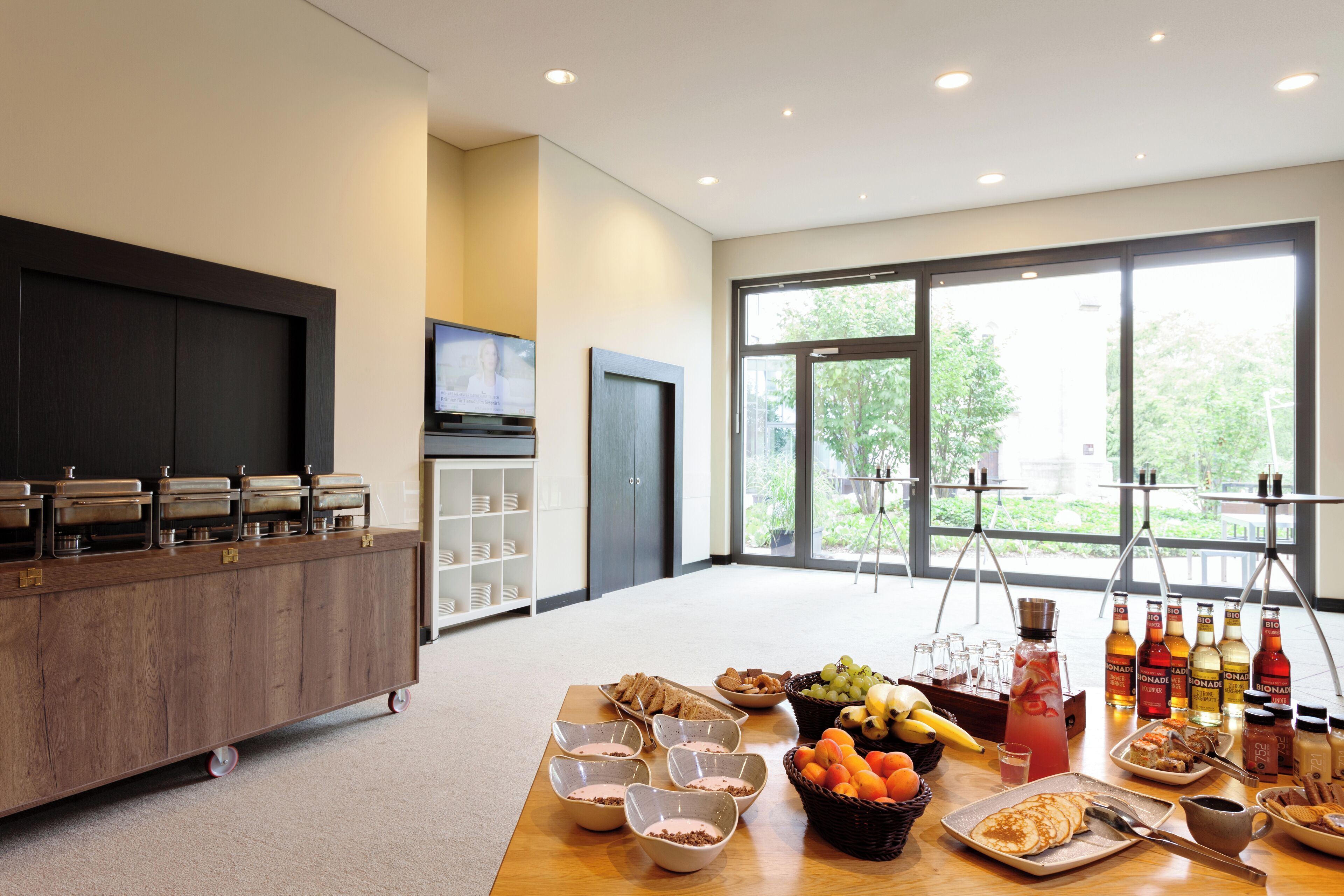 A breakfast buffet with various food items and beverages, a television, and a large window with a view of greenery.