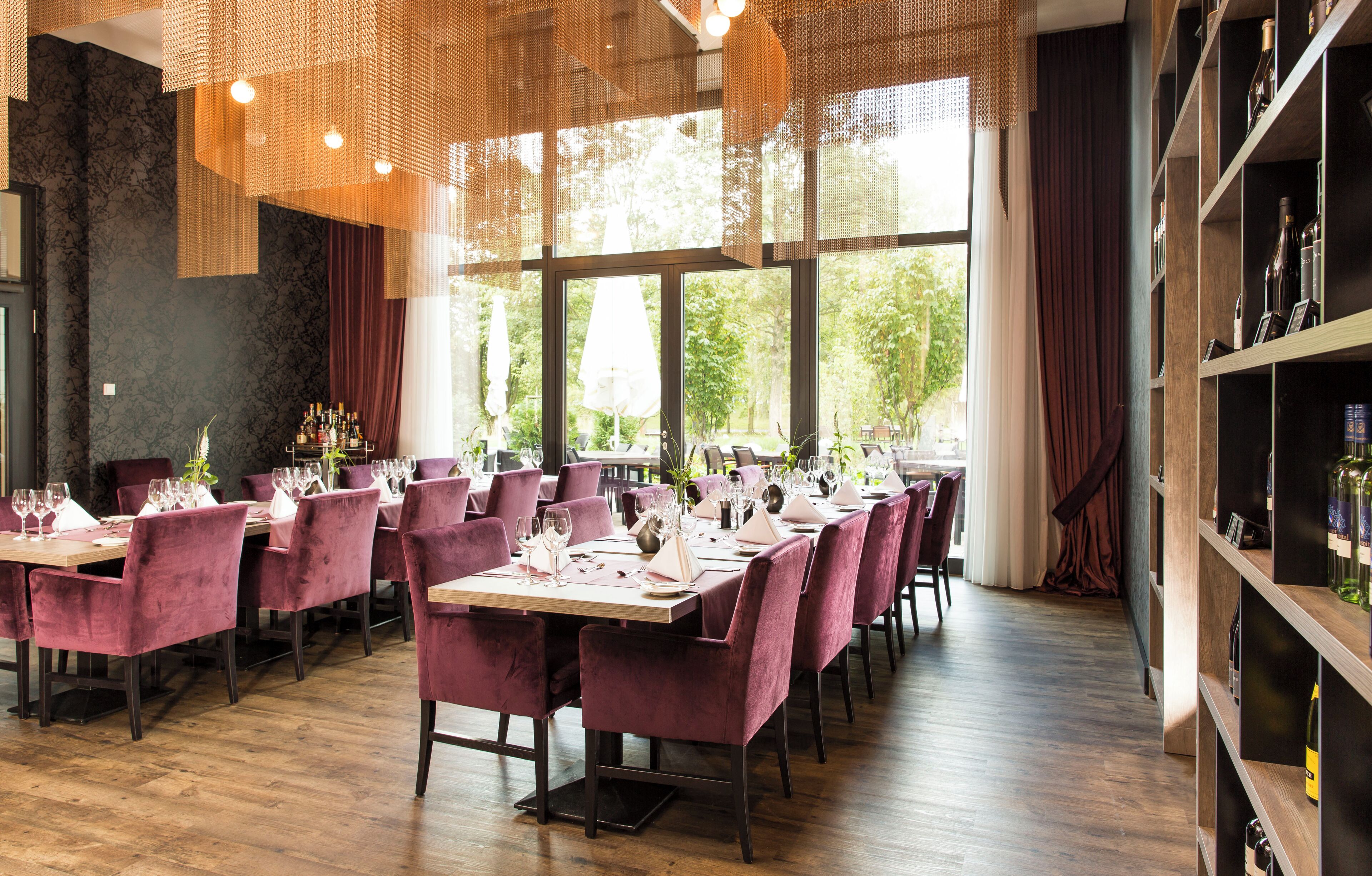 A restaurant with wooden tables and chairs, large windows, and a bookshelf with bottles.