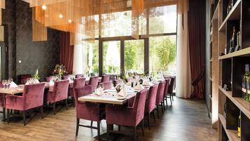 A restaurant with wooden tables and chairs, large windows, and a bookshelf with bottles.
