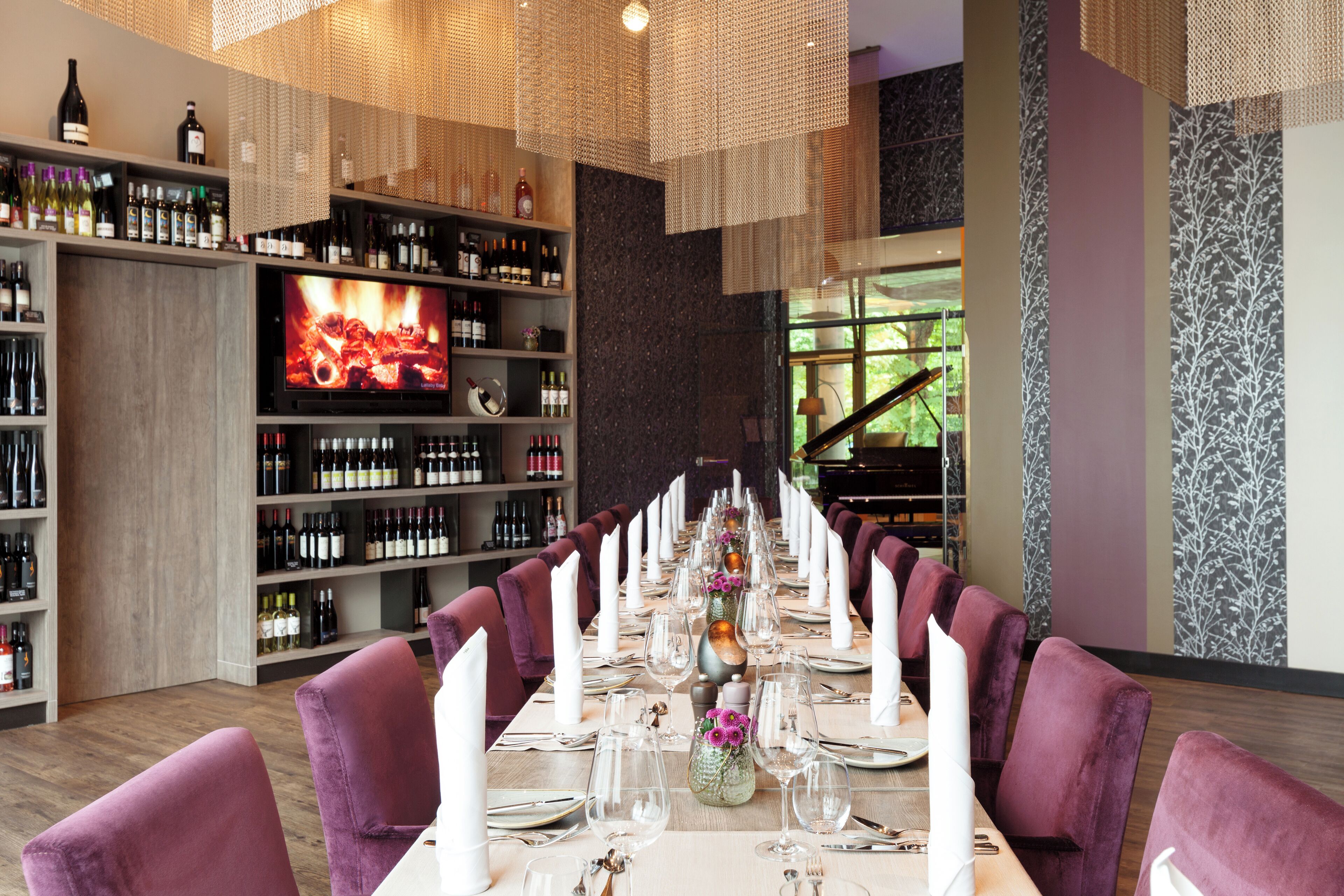 A restaurant with a long dining table set for a formal meal, surrounded by purple chairs, and a wine rack with bottles in the background.