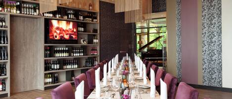 A restaurant with a long dining table set for a formal meal, surrounded by purple chairs, and a wine rack with bottles in the background.