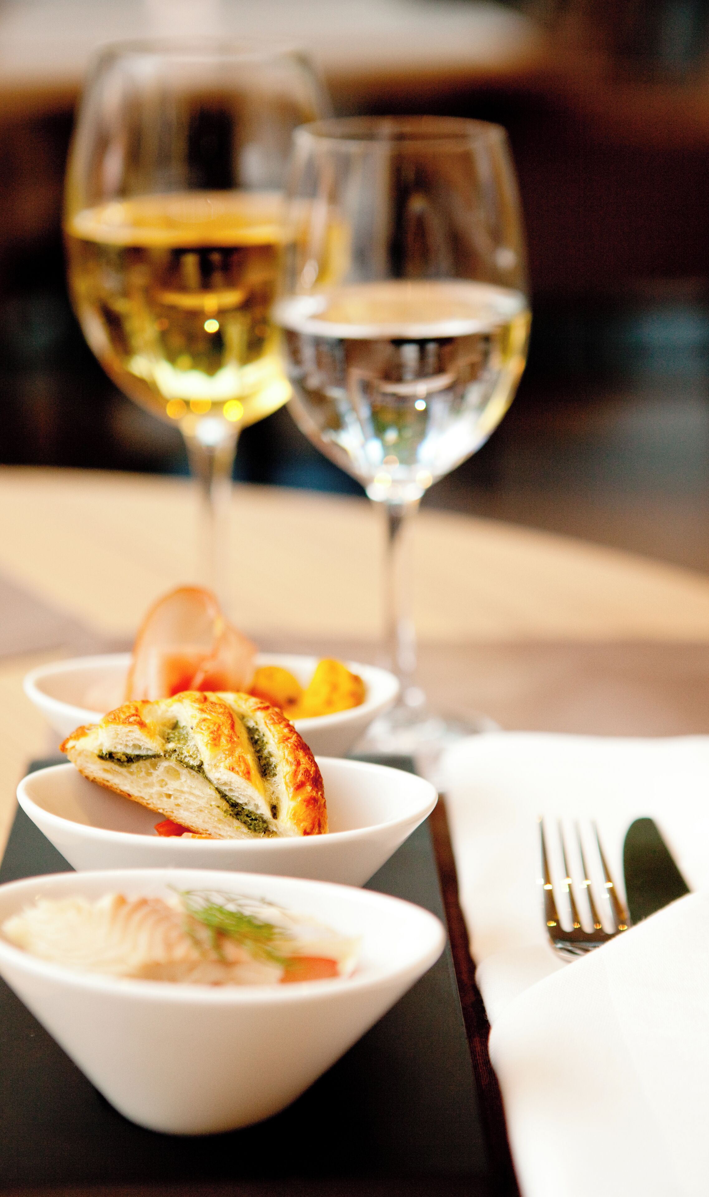 A dining table with three bowls of food, two glasses of white wine, and a fork on a napkin.