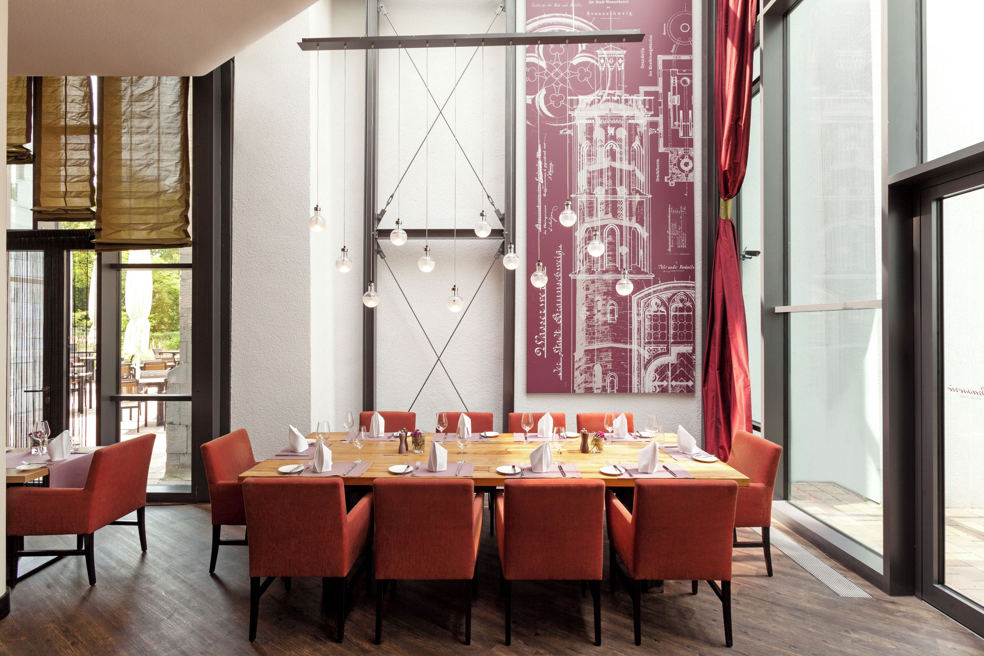A dining area with a long table set for six, surrounded by red chairs, with hanging light fixtures and a large architectural poster on the wall.