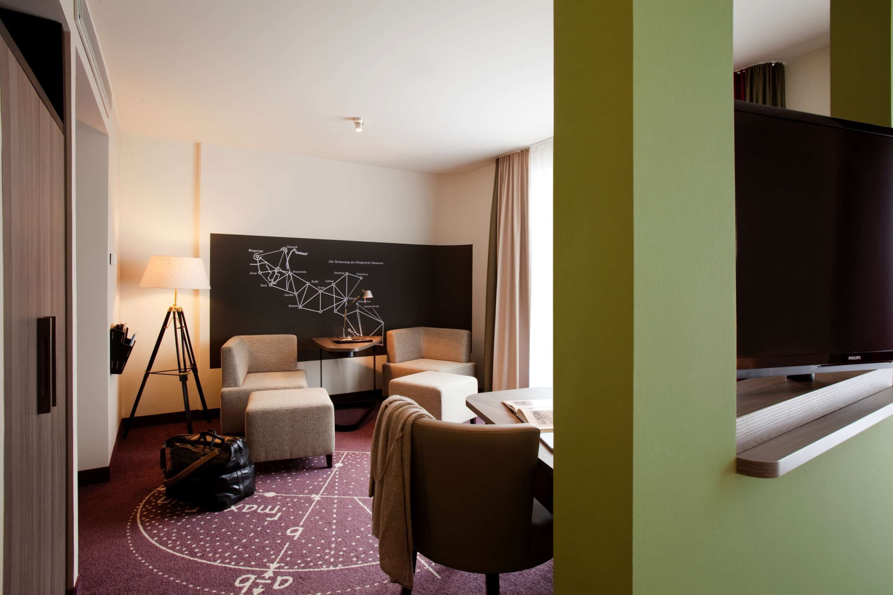 A hotel room with a sofa, armchair, and a desk with a lamp. There is a large TV on the right side.