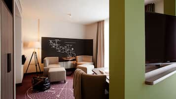 Ein Hotelzimmer mit einer Couch, einem Sessel und einem Schreibtisch mit Lampe. Eine große Fernseher befindet sich auf der rechten Seite.