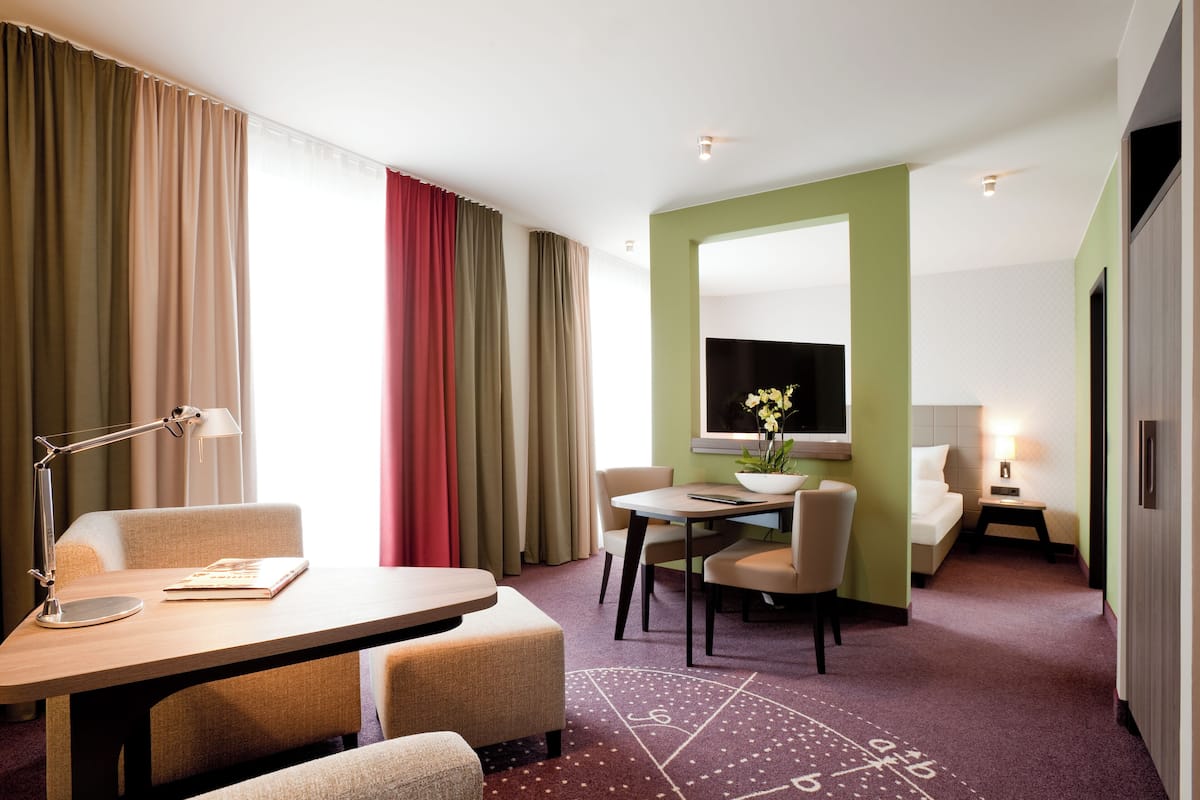 Ein Hotelzimmer mit einem Bett, einem Schreibtisch, einem Stuhl, einer Lampe, einem kleinen Tisch mit Pflanze und einem Fenster mit Vorhängen.