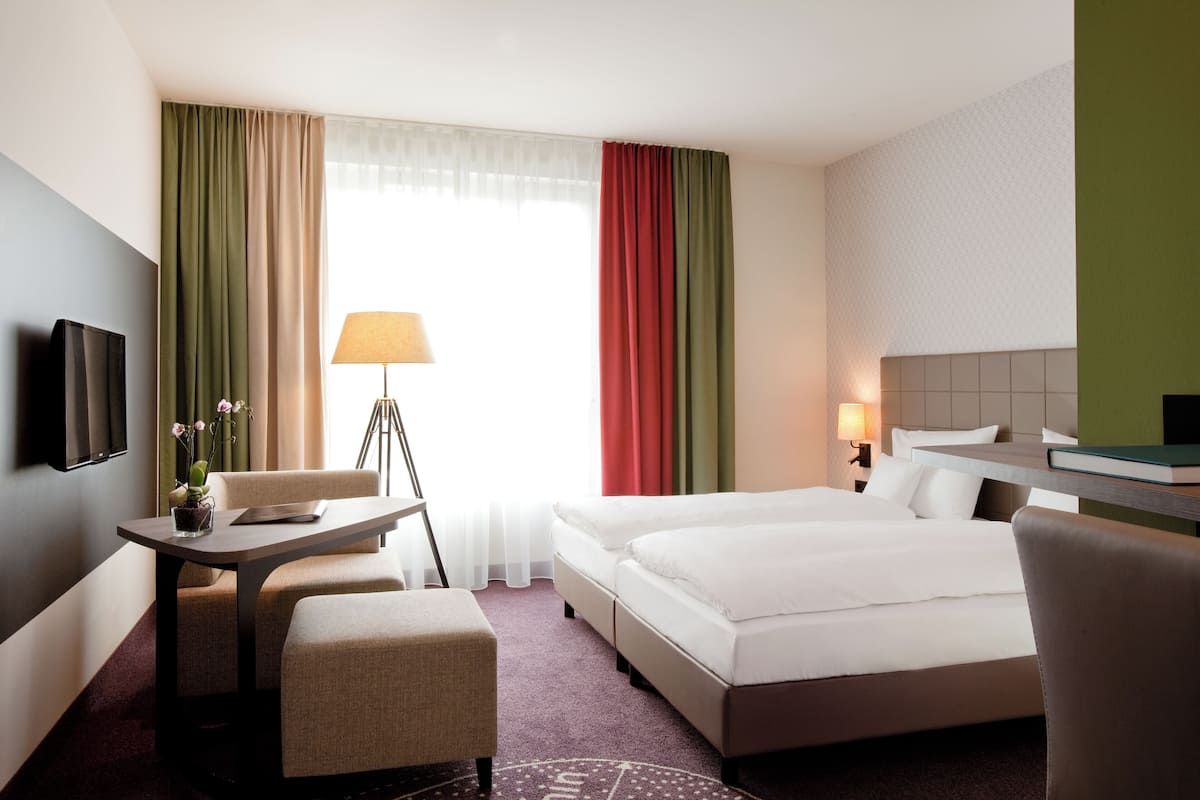 Ein Hotelzimmer mit Bett, Schreibtisch mit Lampe, Stuhl, Fernseher und Fenster mit Vorhängen.