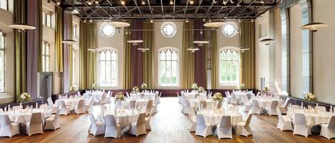 A large hall with multiple round tables set for a formal event, featuring a high ceiling with exposed beams and large windows.