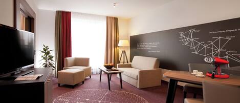 A hotel room with a TV, two armchairs, a sofa, a coffee table, a lamp, and a map on the wall.