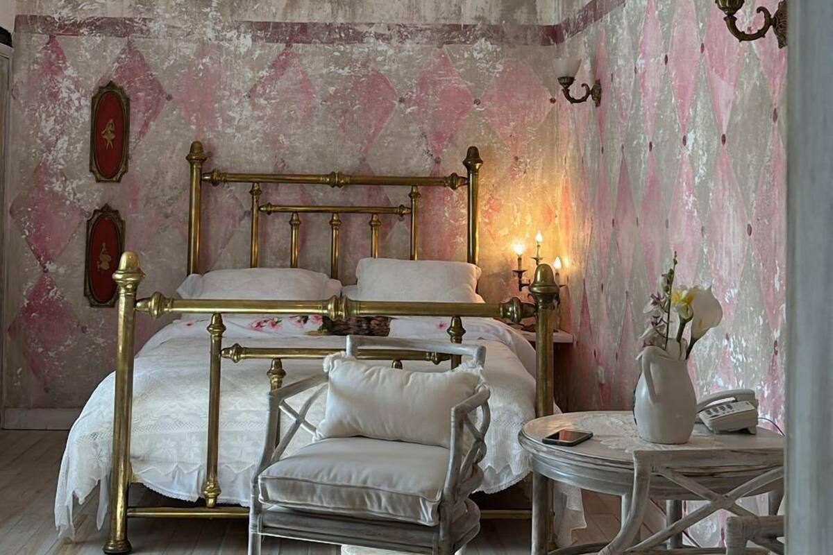 A bedroom with a large bed, a chandelier, a vintage chair, and a small table with a vase and a phone.