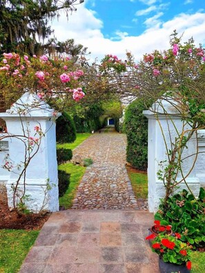 A stone pathway leading through a garden with white pillars and pink flowers. - Casa de Hacienda Su Merced (Puembo)