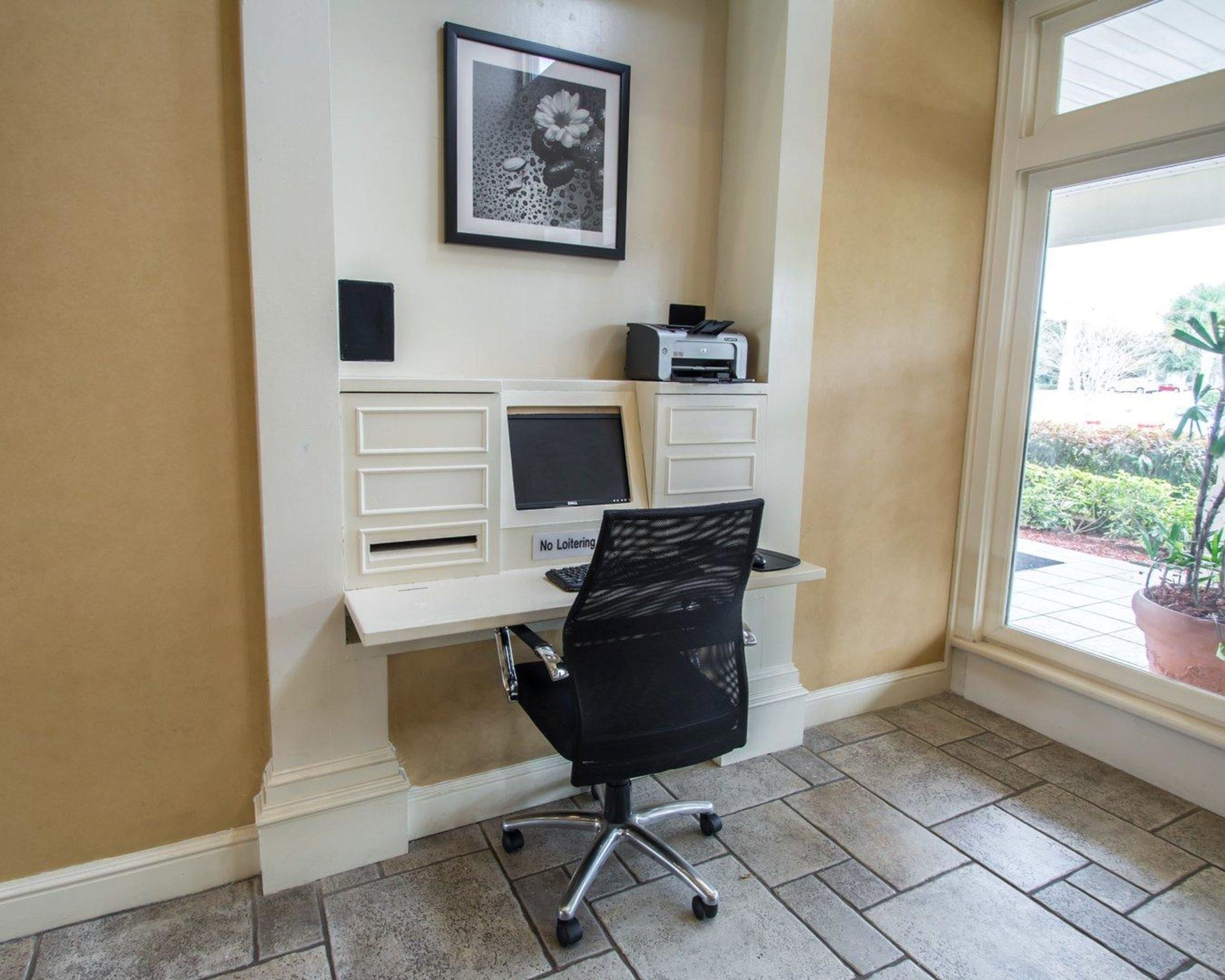Un petit espace de bureau comprenant un bureau, un ordinateur et une chaise. Un tableau encadré est accroché au mur, et une imprimante se trouve sur le bureau.