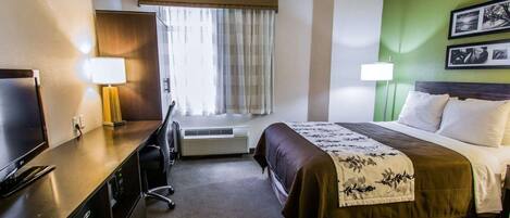 A hotel room with a bed, a desk with a TV, a lamp, a window with curtains, an air conditioning unit, and a green accent wall.