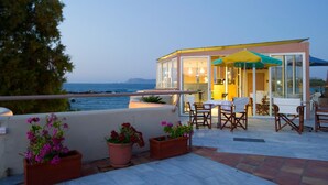 Una terraza con mesas al aire libre, una estructura cubierta de cristal con un paraguas amarillo y vista al mar.