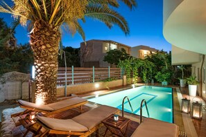 A swimming pool with lounge chairs, a palm tree, and a modern building in the background. - Seafalios Apartments (Chania)
