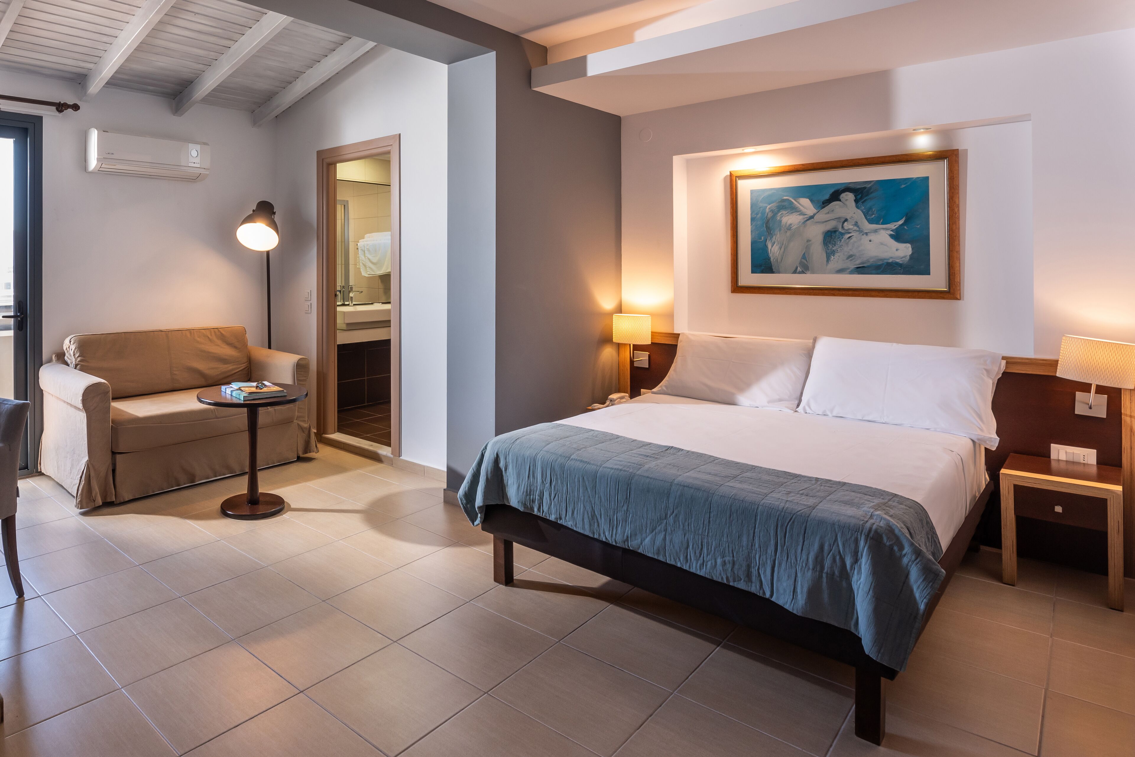 A hotel room with a bed, a sofa, a small table, a lamp, and a painting on the wall.