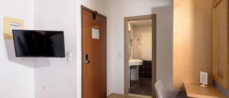 A hotel room with a bed, a desk, a chair, a television, and a bathroom visible through an open door.
