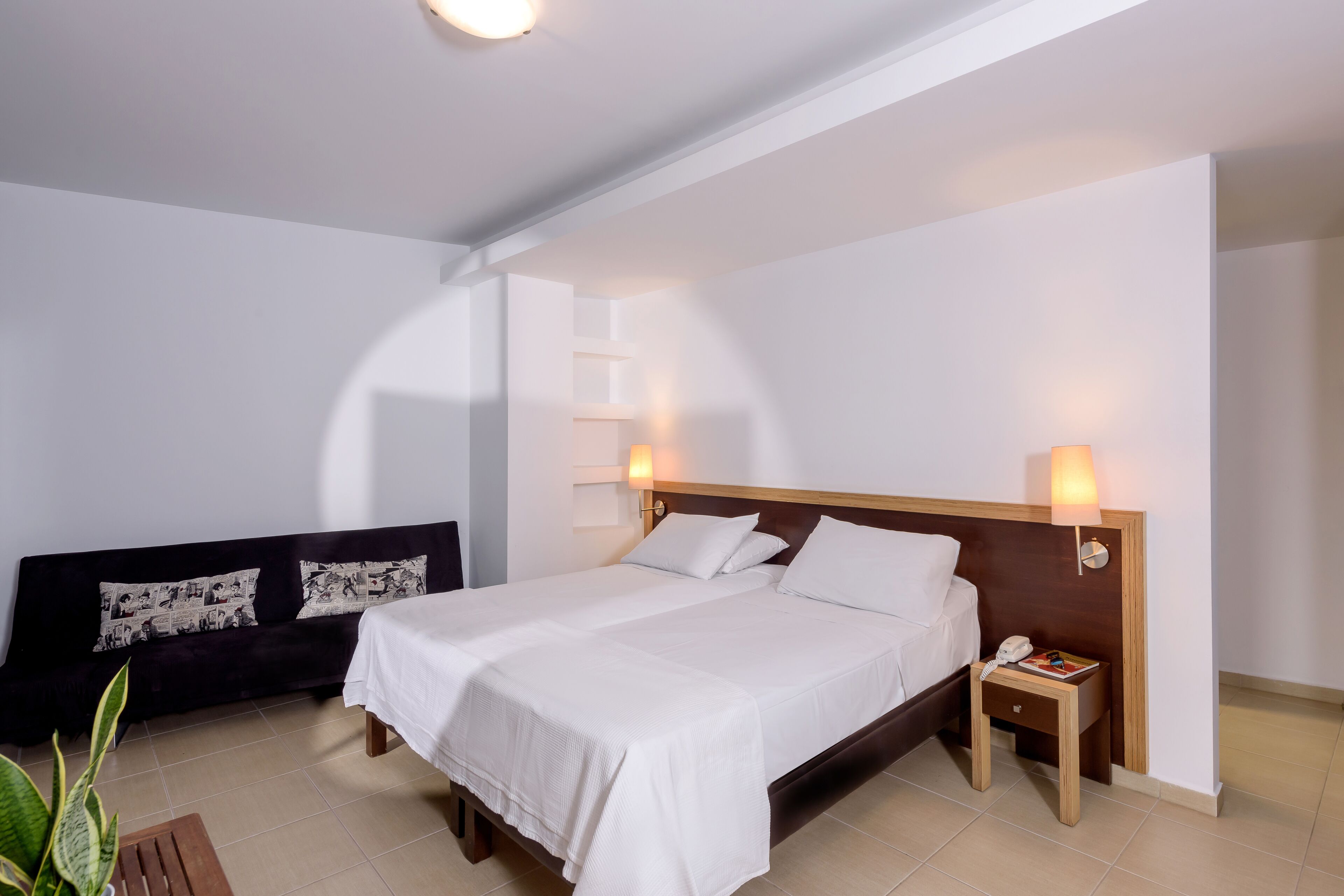 Quarto de hotel com uma cama grande, mesas de cabeceira, um sofá e uma estante fixada na parede.
