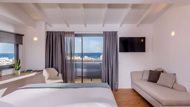 A modern hotel room with a large bed, a sofa, a TV mounted on the wall, and a balcony with a view of the city. - Iraklion Hotel (Heraklion)