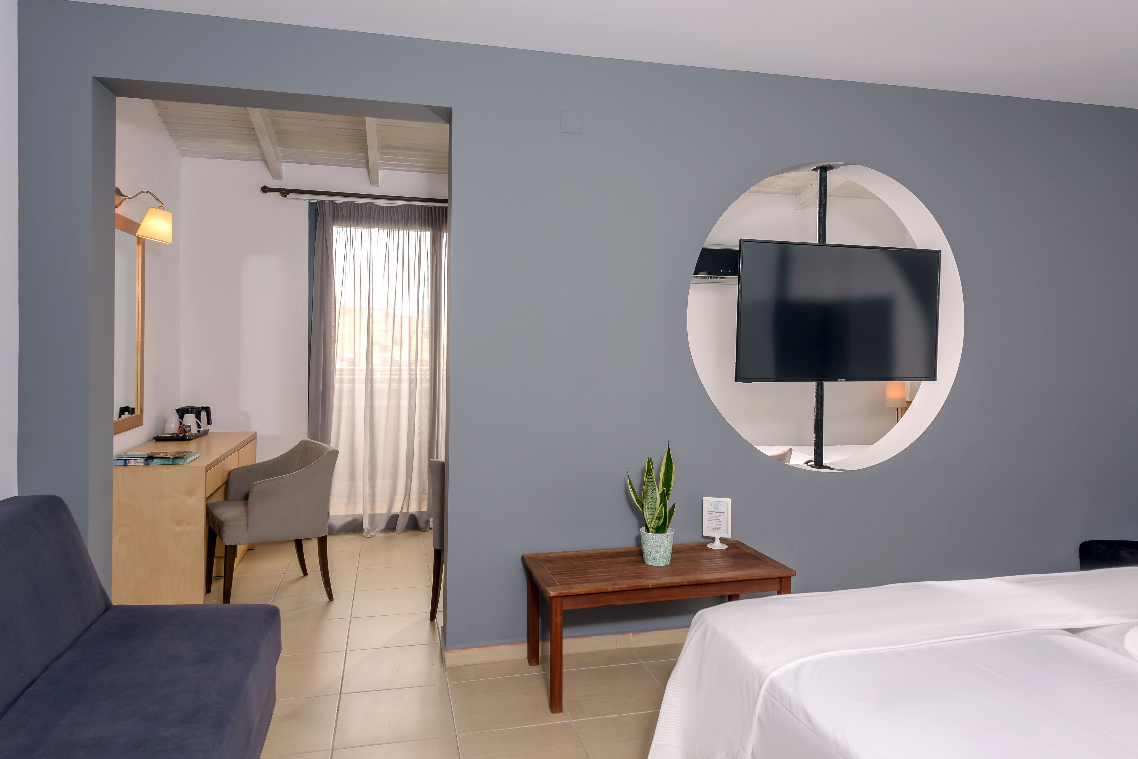 A hotel room with a bed, a desk with a chair, a small table with a plant, a round mirror, and a TV mounted on the wall.