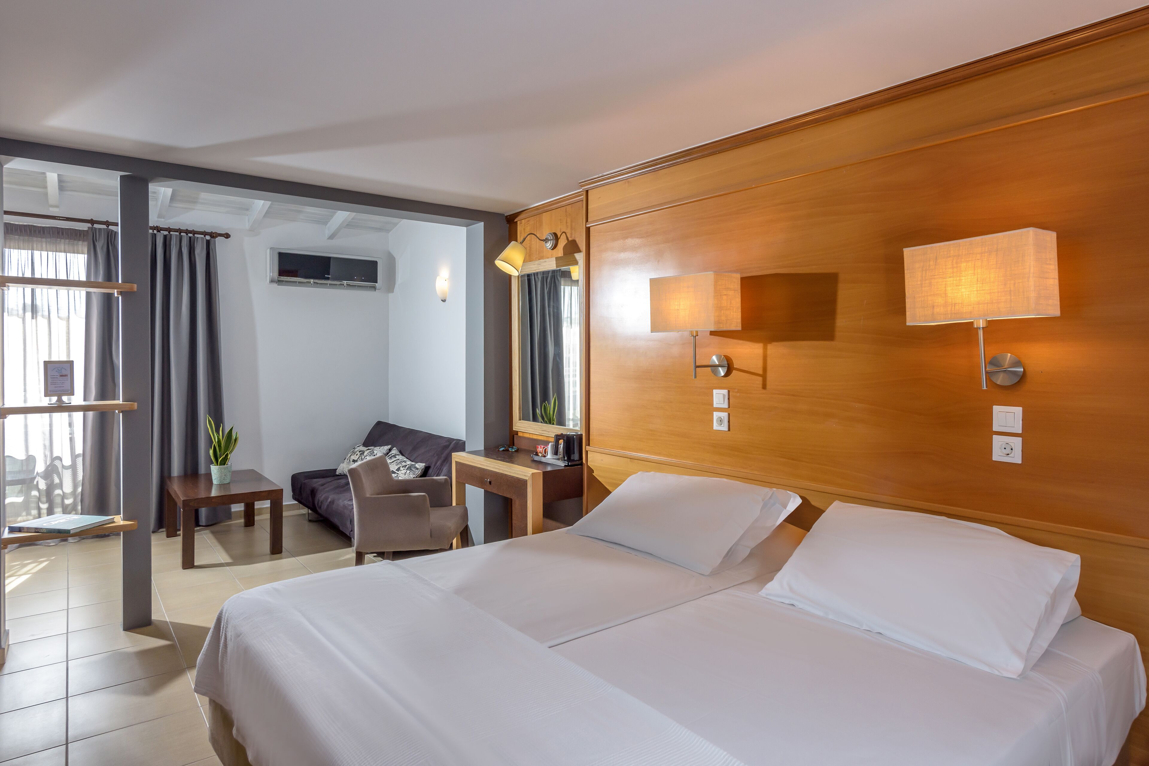 A hotel room with a large bed, a desk, a chair, a small table, a potted plant, and a window with curtains.