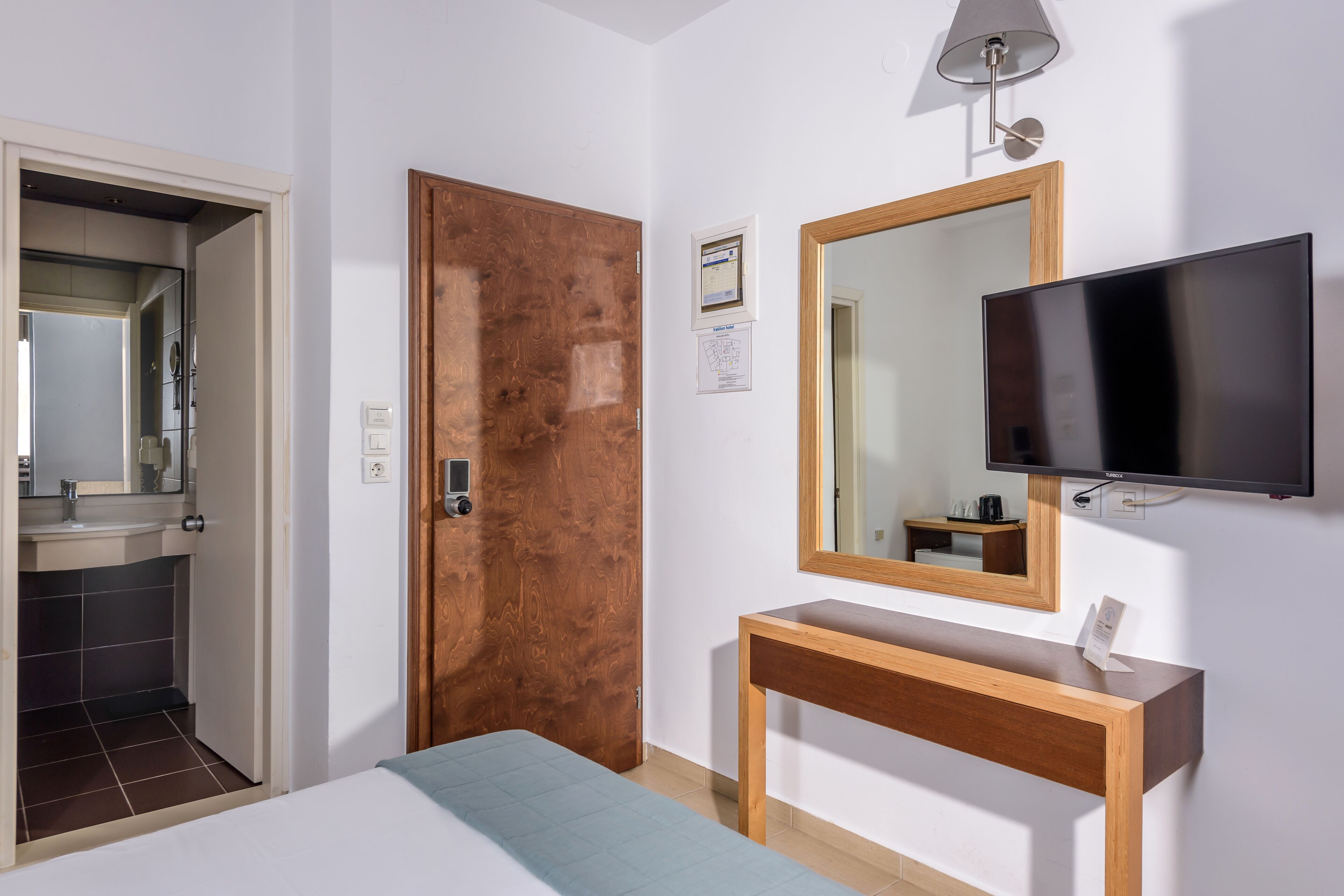 A hotel room with a bed, a wooden door, a bathroom with a mirror, a TV mounted on the wall, and a small table.