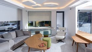 A modern interior with a sofa, chairs, a wooden table, and a large window with a view of the street. - Iraklion Hotel (Heraklion)