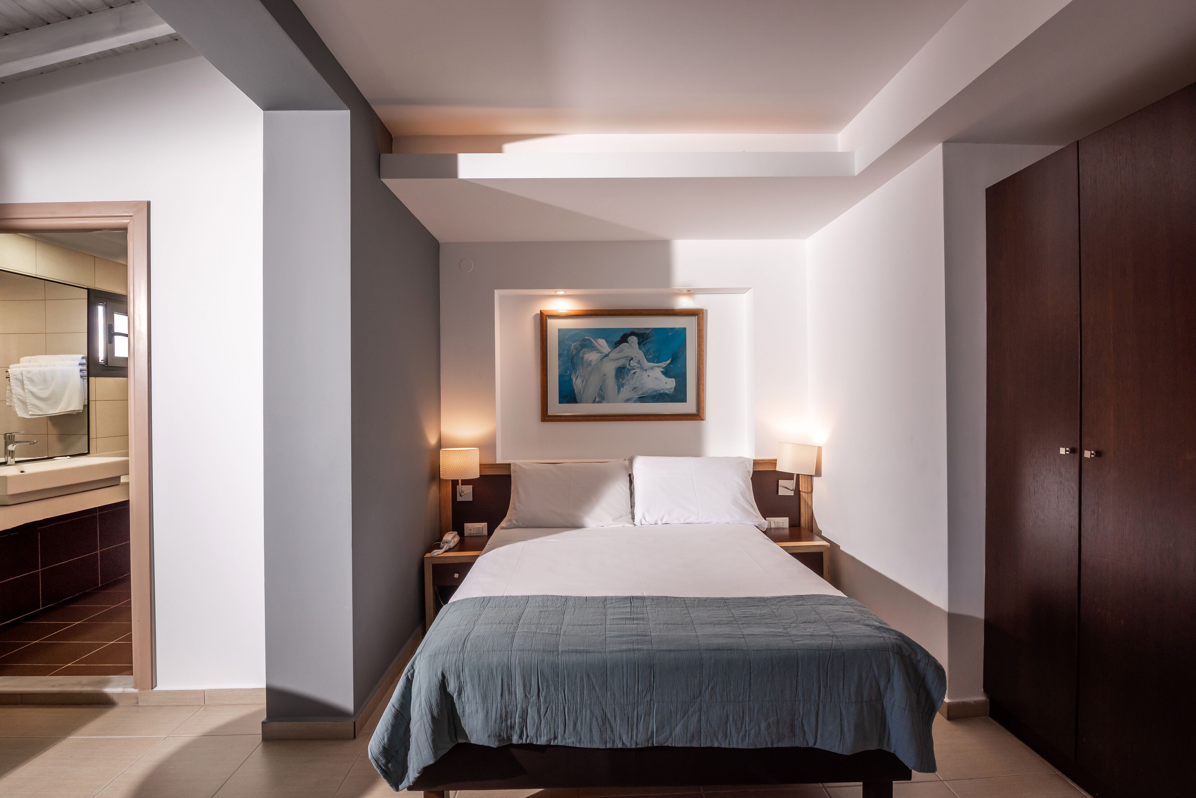 Una camera d'albergo con un letto, comodini, un quadro sopra il letto, un armadio e un bagno parzialmente visibile attraverso una porta aperta.