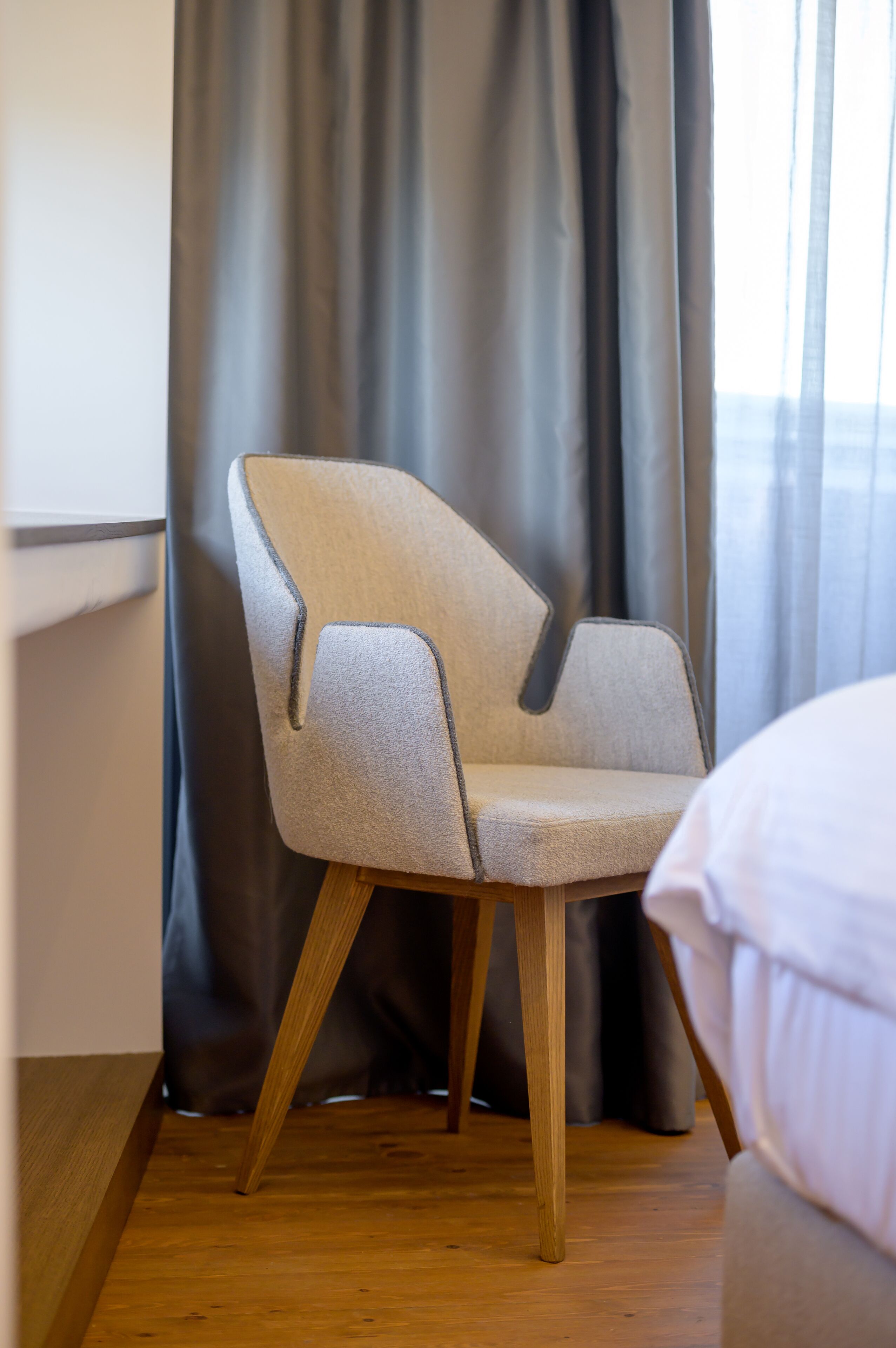Un fauteuil moderne doté d’un piétement en bois et d’un siège recouvert d’un tissu de couleur claire, placé devant une fenêtre ornée de rideaux gris.