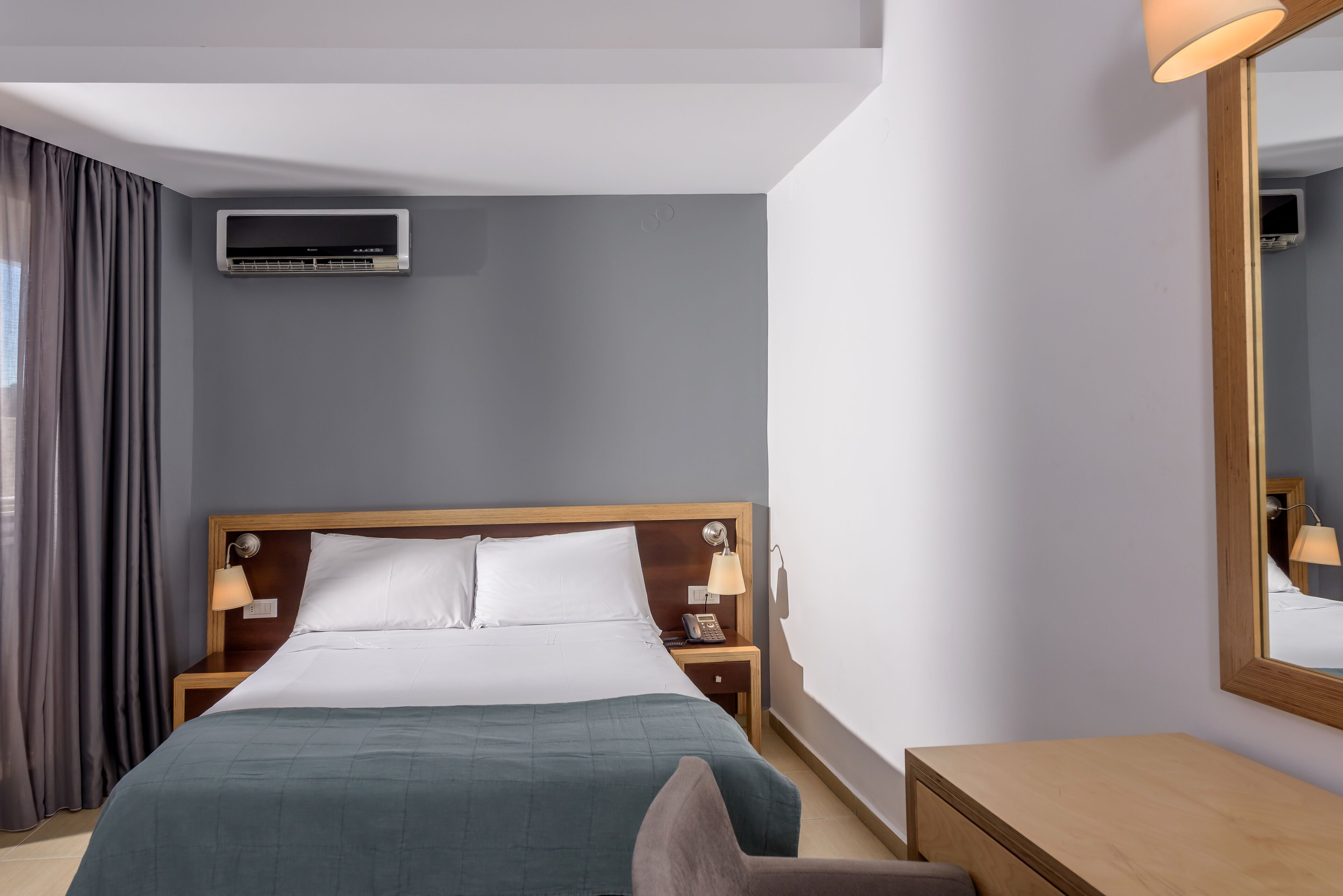 A hotel room with a bed, bedside lamps, a nightstand, a wooden desk, a mirror, and an air conditioning unit.