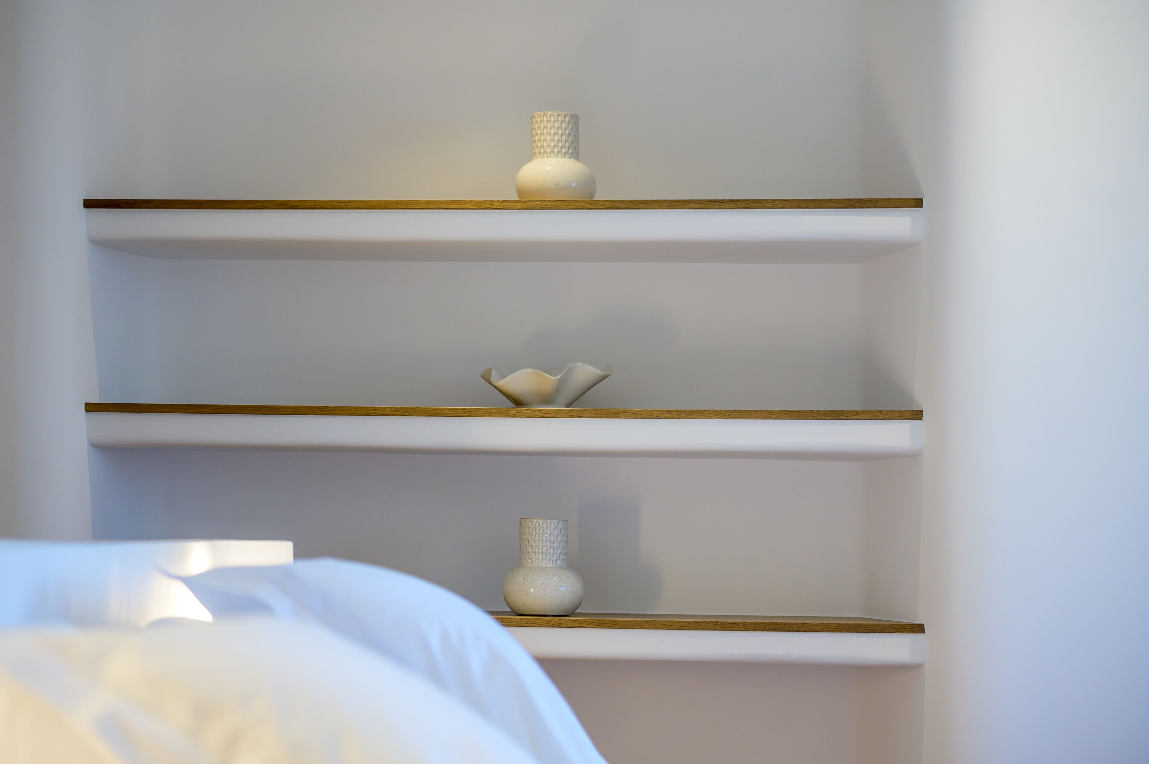Une chambre à coucher avec un lit, des murs blancs et trois étagères ornées d’objets décoratifs.
